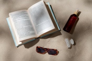 leisure and summer holidays concept - open book, sunscreen, hair clip and sunglasses on beach sand. book, sunglasses and sunscreen on beach sand