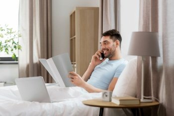people, technology and remote job concept - man with folder and laptop computer calling on smartphone in bed at home bedroom. man with folder calling on phone in bed at home