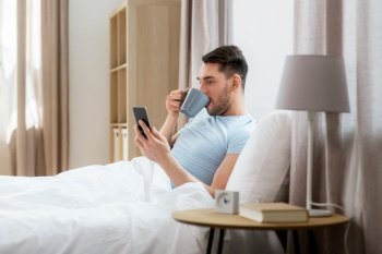 people, bedtime and rest concept - happy smiling man with smartphone drinking coffee in bed at home. happy man with smartphone in bed at home