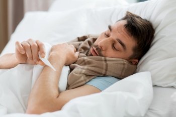 people, health and fever concept - sick man in scarf measuring temperature by thermometer lying in bed at home. sick man measuring temperature by thermometer