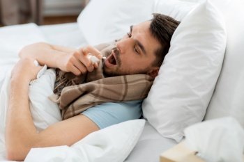 people and health problem concept - unhappy sick man spraying his throat with oral spray lying in bed at home. sick man with oral spray lying in bed at home