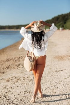 people, summer holidays and leisure concept - woman in white shirt and straw hat with bag walking along beach. woman with bag walking along summer beach