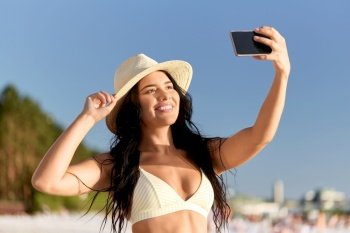 people, summer and swimwear concept - happy smiling young woman in bikini swimsuit and straw hat taking selfie with smartphone on beach. smiling woman in bikini taking selfie on beach