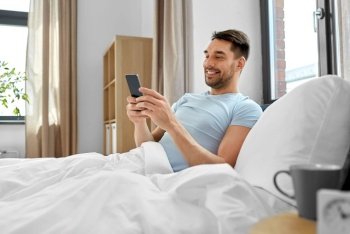 technology, internet and people concept - smiling man texting on smartphone in bed at home in morning. man with smartphone in bed in morning at home