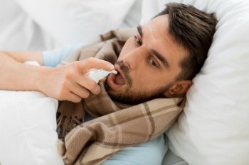 people and health problem concept - unhappy sick man spraying his throat with oral spray lying in bed at home. sick man with oral spray lying in bed at home