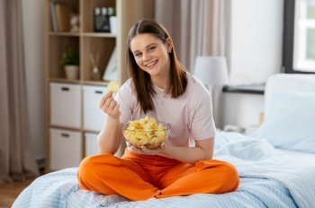 fast food and people concept - happy girl eating crisps sitting on bed at home. happy girl eating crisps sitting on bed at home