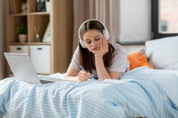 school, online education and e-learning concept - bored teenage student girl in headphones with laptop computer writing to notebook lying on bed at home. student girl in headphones learning at home