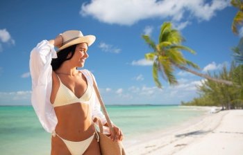 travel, tourism and summer holidays concept - portrait of young woman in bikini swimsuit, white shirt and straw hat with bag over tropical beach background in french polynesia. portrait of woman in bikini and hat on beach