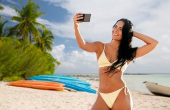 travel, tourism and summer holidays concept - happy smiling young woman in bikini swimsuit taking selfie with smartphone over tropical beach background in french polynesia. smiling woman in bikini taking selfie on beach
