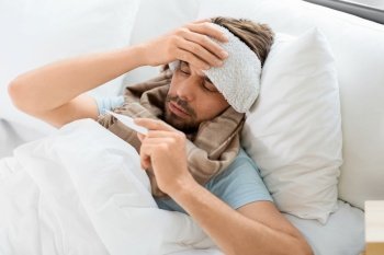 people, health and fever concept - sick man with cold compress on his forehead measuring temperature by thermometer lying in bed at home. sick man measuring temperature by thermometer