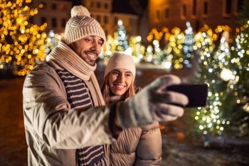 winter holidays and people concept - happy smiling couple taking selfie with smartphone over christmas tree lights in evening city. happy couple taking selfie over christmas lights