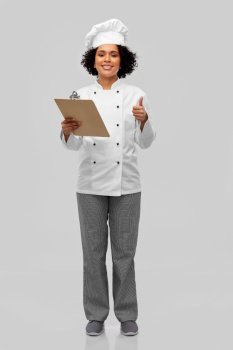 cooking, culinary and people concept - happy smiling female chef in toque and jacket with clipboard showing thumbs up over grey background. female chef with clipboard showing thumbs up