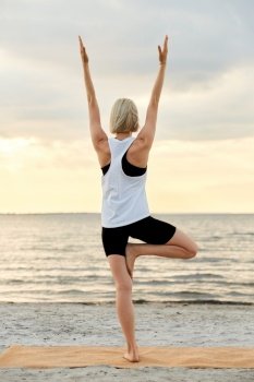 fitness, sport, and healthy lifestyle concept - woman doing yoga tree pose on beach over sunset. woman doing yoga tree pose on beach over sunset