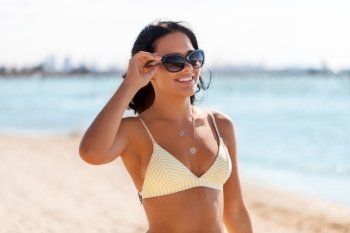 people, summer and swimwear concept - happy smiling young woman in bikini swimsuit and sunglasses on beach. smiling young woman in bikini swimsuit on beach