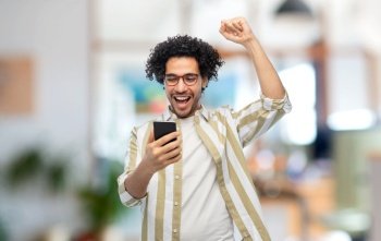 technology, communication and people concept - happy smiling young man in glasses with smartphone celebrating success over office background. happy smiling young man with smartphone at office