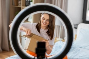 blogging, technology and people concept - happy smiling girl blogger with ring light and smartphone unpacking parcel box at home. happy girl blogger unpacking parcel box at home