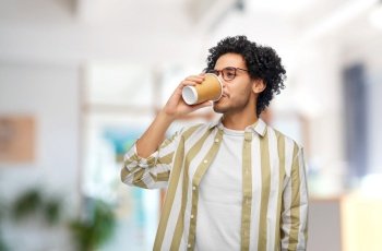 drinks and people concept - young man in glasses drinking takeaway coffee from paper cup over office background. man drinking takeaway coffee at office