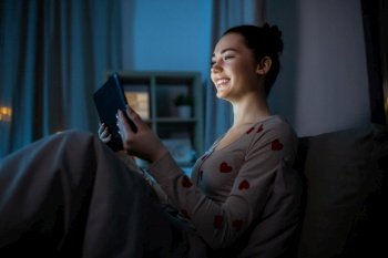 technology, bedtime and people concept - happy smiling teenage girl tablet pc computer sitting in bed at home at night. teenage girl with tablet pc in bed at night