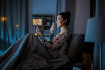technology, bedtime and people concept - happy smiling teenage girl with smartphone and earphones having video call sitting in bed at home at night. teenage girl with phone pc has video call in bed