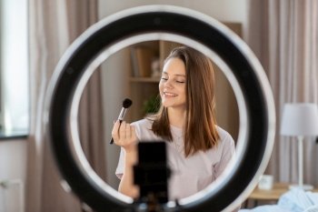 beauty blogging, technology and people concept - happy smiling girl blogger with ring light and smartphone applying make up at home. girl blogger with ring light applying make up
