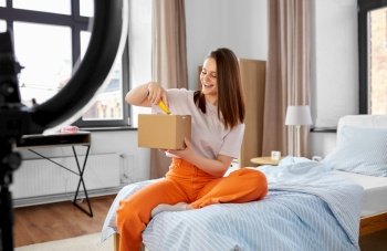 blogging, technology and people concept - happy smiling girl blogger with ring light and smartphone unpacking parcel box with knife at home. happy girl blogger unpacking parcel box at home