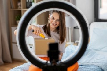blogging, technology and people concept - happy smiling girl blogger with ring light and smartphone unpacking parcel box with knife at home. happy girl blogger unpacking parcel box at home