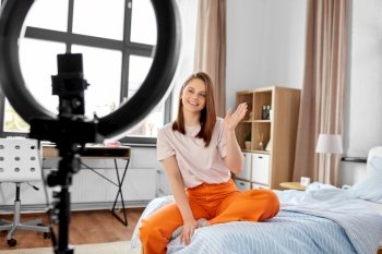 blogging, technology and people concept - happy smiling girl blogger with ring light and smartphone waving hand at home. girl blogger with ring light streaming at home