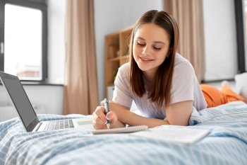 school, online education and e-learning concept - teenage student girl with laptop computer writing to notebook lying on bed at home. teenage student girl writing to notebook at home