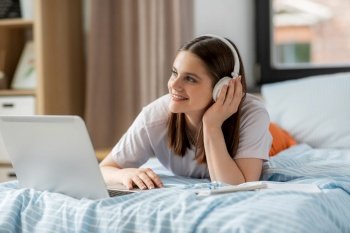 school, online education and e-learning concept - happy smiling teenage student girl in headphones with laptop computer lying on bed and listening to music at home. happy student girl in headphones learning at home