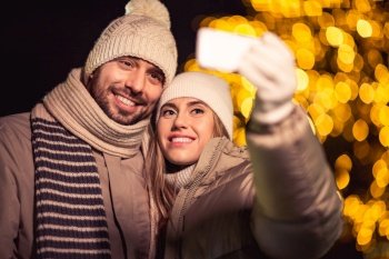 winter holidays and people concept - happy smiling couple taking selfie with smartphone over christmas lights in evening. happy couple taking selfie over christmas lights