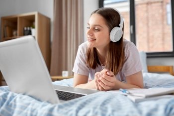 school, online education and e-learning concept - happy smiling teenage student girl in headphones with laptop computer lying on bed at home. happy student girl in headphones learning at home
