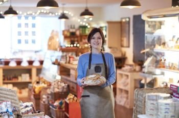 Portrait Of Smiling Female Owner Of Delicatessen Shop Wearing Apron Holding Loaf Of Bread