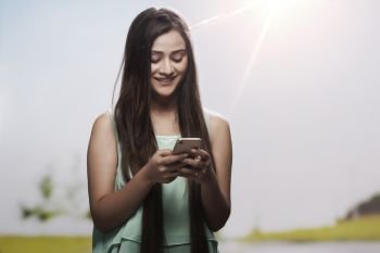 Portrait of a smiling teenage girl using her mobile phone outdoors with sun in the background