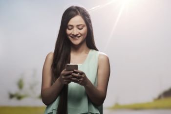 Portrait of a smiling teenage girl using her mobile phone outdoors with sun in the background
