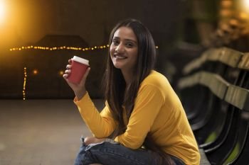 Portrait of happy young woman sitting on bench holding coffee cup on street