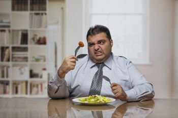 Overweight man sitting at dining table with plate full of vegetable salad holding a tomato slice with fork and looking at it with dislike