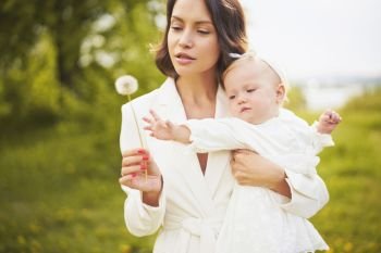 Outdoor fashion portrait of young beautiful mother and little cute daughter blowing to dandelion on green meadow. Spring image