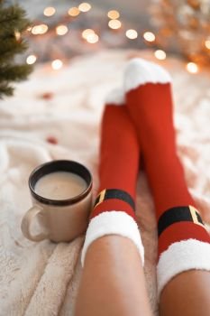 Lifestyle home photo of feet in Santa’s socks near the Christmas tree. Woman sitting at the blanket, drinks hot beverage and relaxes  warming up their feet in woollen socks. Winter and Christmas holidays concept