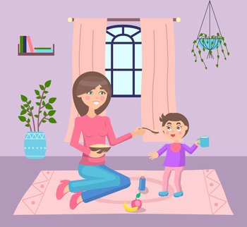 Young mother feeding her son, sit at floor near standing kid. Little boy holding cup and smiling. Lunch, dinner or supper time. Woman give a spoon with meal to child. Apple and banana at floor. Mother feeding her son sitting at carpet at floor near standing kid with cup in hand smiling
