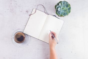 Open empty notebook on white wooden table with cup of coffee, succulent flower and someones hand with pen. Open empty notebook
