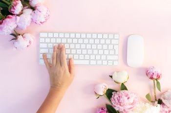 Flat lay home office workspace - modern keyboard with peony flowers and someones one hand typing, copy space on pink background. Top view home office workspace