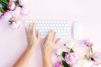 Flat lay home office workspace - modern keyboard with peony flowers and someones hands typing, copy space on pink background. Top view home office workspace