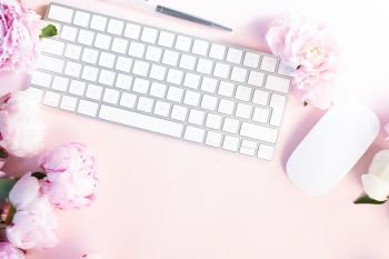 Flat lay home office workspace - modern keyboard with female accessories and peony flowers, copy space on pink desk background. Top view home office workspace