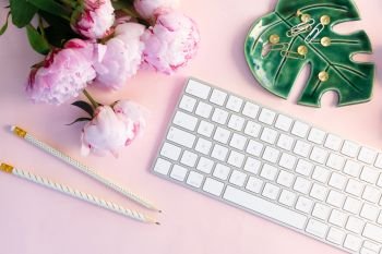 Flat lay top view home office workspace - modern keyboard with female accessories and peony flowers, copy space on pink background. Top view home office workspace