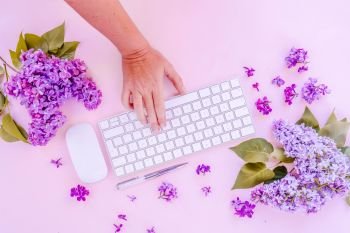 Flat lay home office workspace - modern keyboard with someone hand typing, fresh lilac flowers, toned. Top view home office workspace