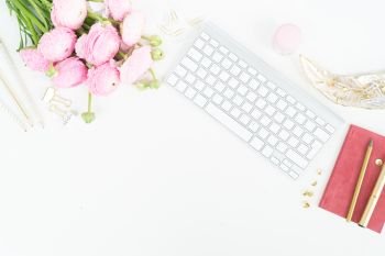 Flat lay home office workspace - modern keyboard with female accessories and ranunculus flowers, copy space on white background. Top view home office workspace