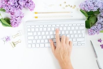 Flat lay top view home office workspace - hand typing on modern keyboard with lilac flowers on white background. Top view home office workspace