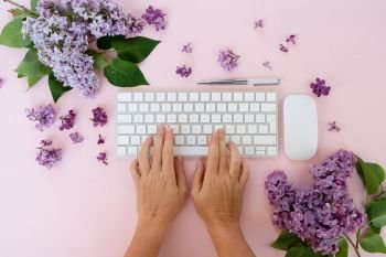 Flat lay home office workspace - modern keyboard with two hands typing, fresh lilac flowers. Top view home office workspace