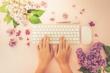 Flat lay home office workspace - modern keyboard with two hands typing, fresh lilac flowers, toned. Top view home office workspace