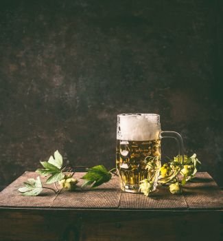 Cold mug of beer with foam on a rustic table with a vine and cones of hops opposite a dark wall, front view.